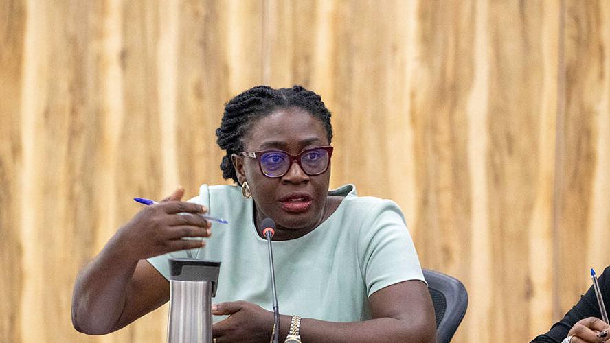 A person seated at a conference table speaks into a microphone while gesturing with one hand. The individual is holding a pen in the other hand. Another person is partially visible on the right, also holding a pen. The background features a wooden panel wall.