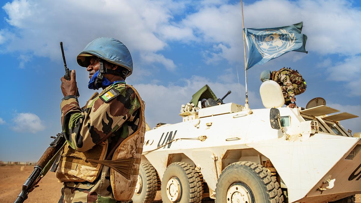 A Nigerian peacekeeper in uniform and a blue helmet talking into a radio holding a gun. In the background is a white tank flying the blue UN flag. They are both in the desert with blue sky with clouds behind them.