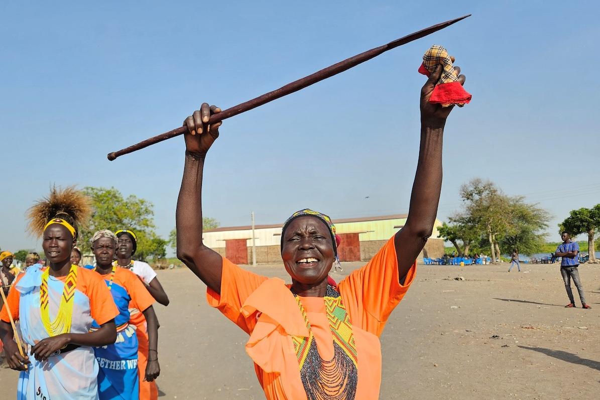 A woman wearing a bright orange t-shirt and large necklace is smiling and holding a wooden object above their head in jubilation. There are some other women in the background. 