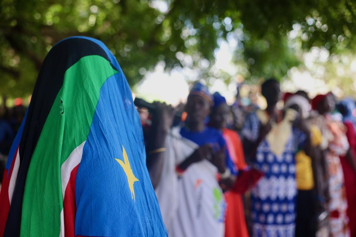 Mental health problems, what causes them and their potentially fatal consequences are difficult but necessary to talk about. During an all-female community dialogue in Kuajok, participants did A person in the foreground is facing away from the camera and has covered their head with the South Sudanese flag. In the background, there are more people in bright clothing which have been blurred. All of the these people are standing under trees.