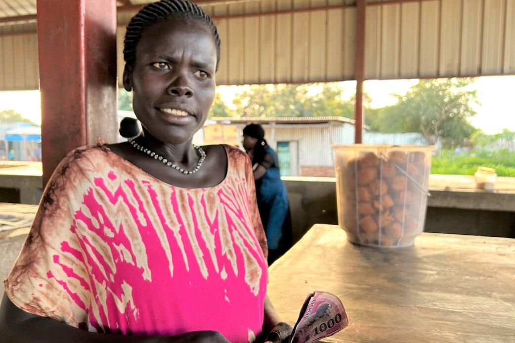 UNMISS' Radio Miraya went live from Aru Junction with stories of how traders and farmers are benefiting from relative stability and peace with the support of peacekeepers from Nepal. Person standing at a market stall holding currency notes, with a container of fried snacks on the counter and open-air structures in the background.