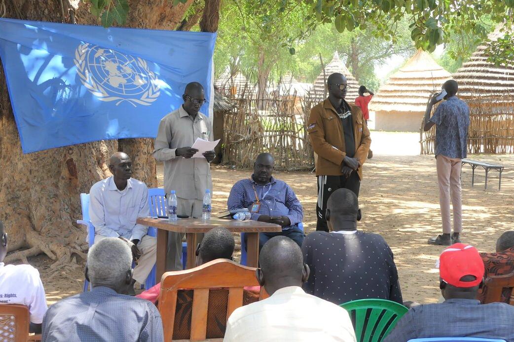 Outdoor meeting under a large tree with a UN flag displayed. Several people are seated on chairs facing a small table where others are standing and speaking. Traditional huts are visible in the background.