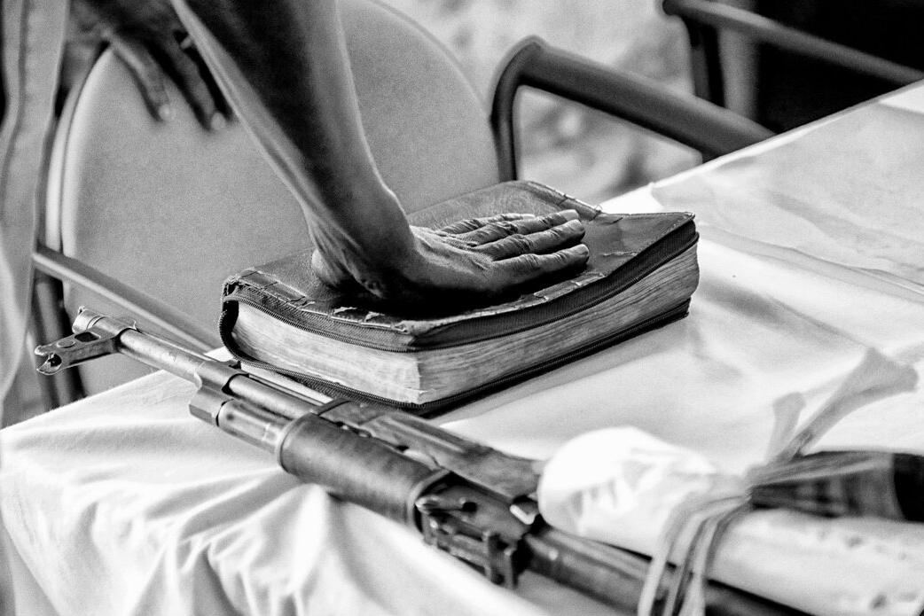 Close-up of a person’s hand resting on a worn book on a table, with a rifle placed beside it.
