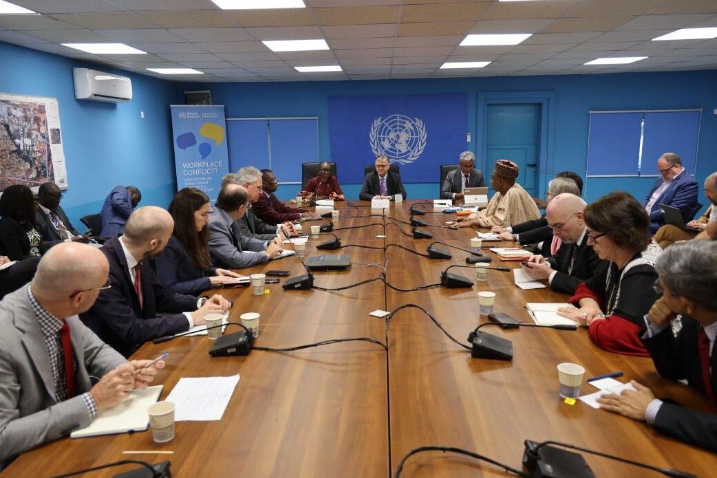 Meeting in a UN conference room with participants seated around a long wooden table, microphones and notebooks in front of them, and a UN emblem on the blue wall