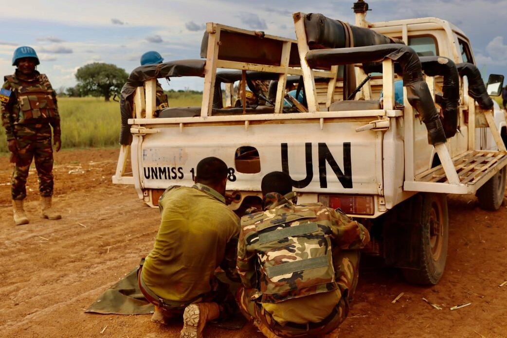 Two uniformed personnel crouch near the rear of a UN-marked pickup truck on a dirt road, while another stands nearby wearing a blue helmet.