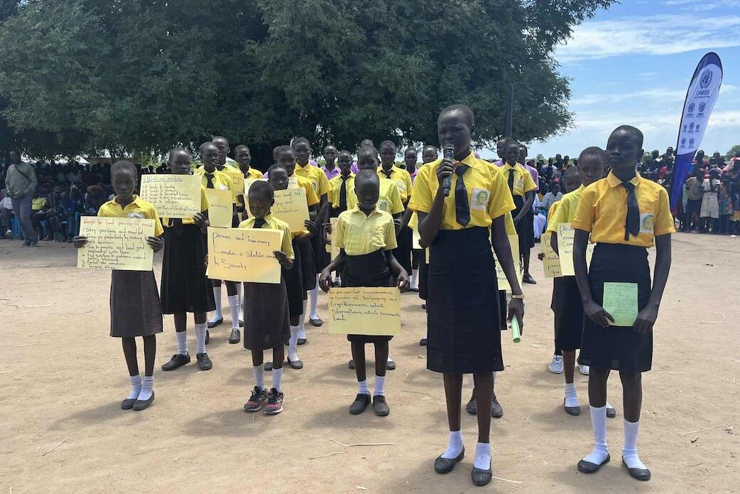Group of schoolchildren in yellow shirts and dark skirts or trousers holding handwritten signs and papers during an outdoor event, with a crowd and banner in the background.