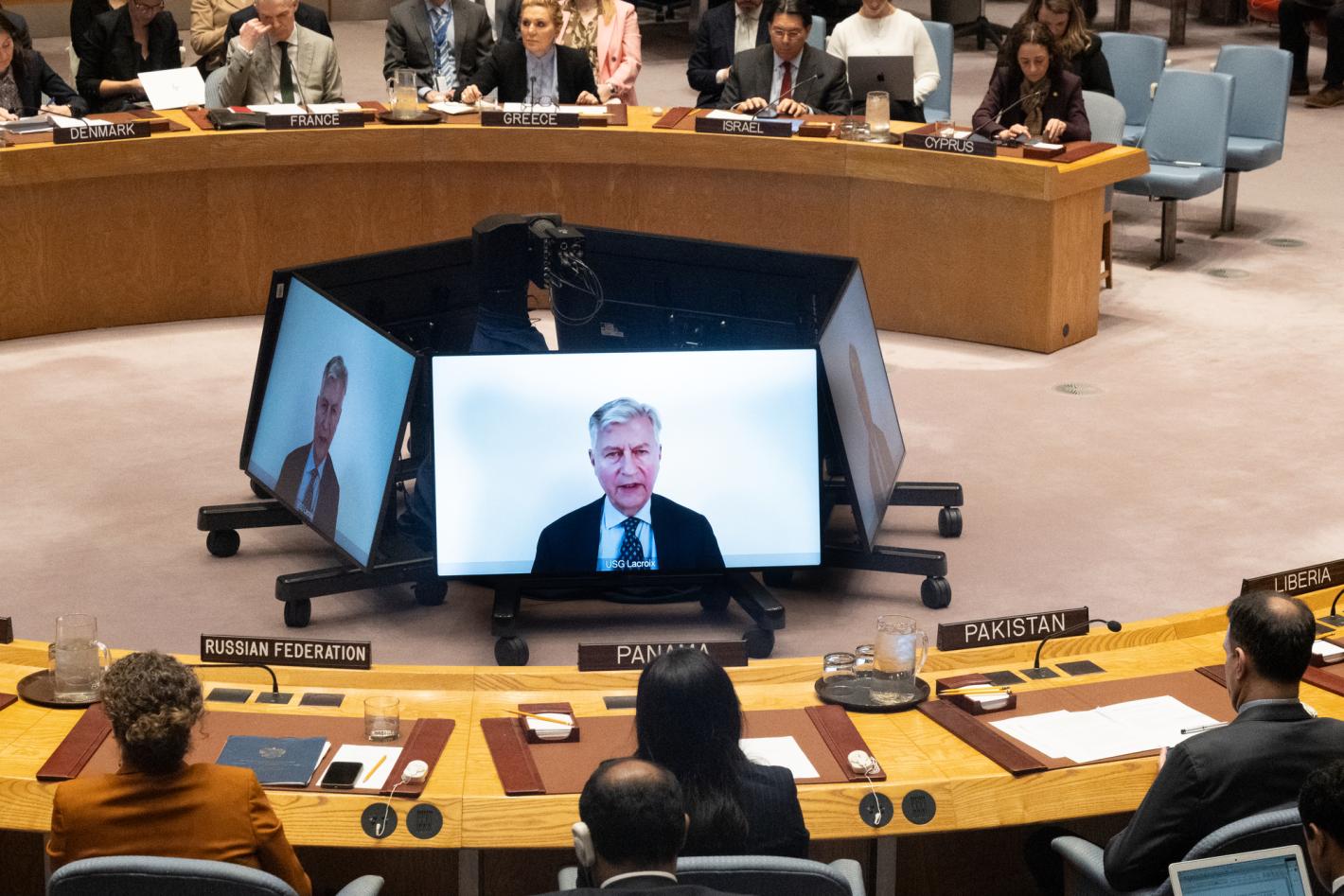 A Security Council meeting room with delegates seated around a circular table, nameplates visible for several countries. At the center, multiple large monitors display a remote briefing speaker. Attendees are focused on the screens, and documents, microphones, and translation earpieces are arranged on the desks.