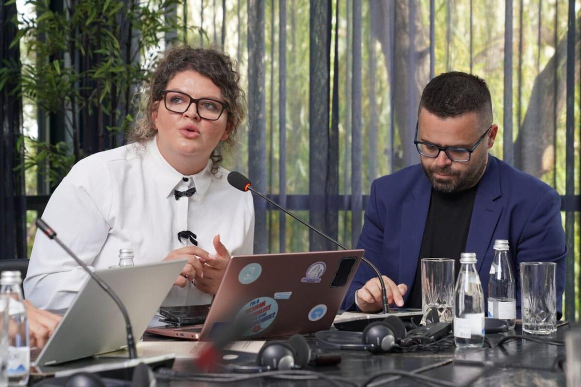 Two people seated at a conference table with laptops, microphones, and water bottles. One laptop has multiple stickers on its cover. The individuals appear to be engaged in conversation or presentation. The background includes large windows with vertical blinds and trees visible outside.