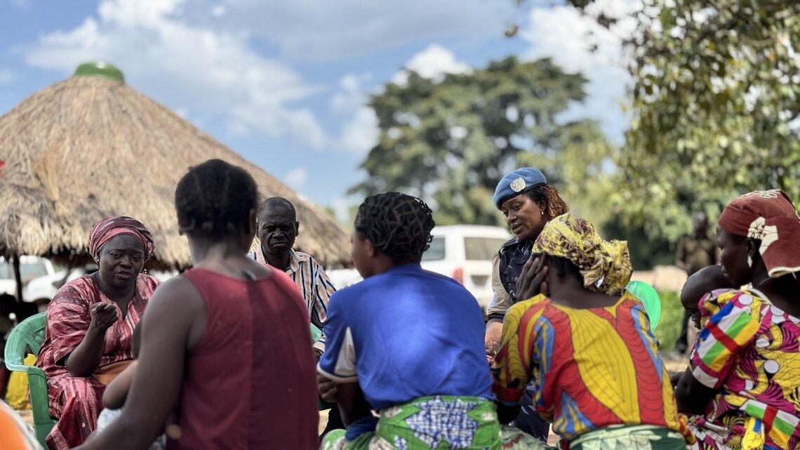 Community members sit outside, speaking with a UN peacekeeper in a blue berret