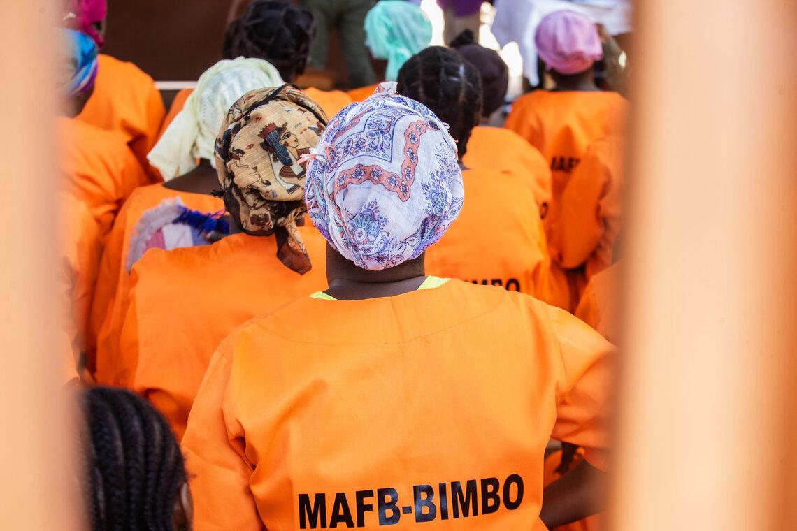 Women wearing bright orange uniforms labeled "MAFB-BIMBO" sit together, viewed from behind, with colorful headscarves in a group setting framed by vertical bars.