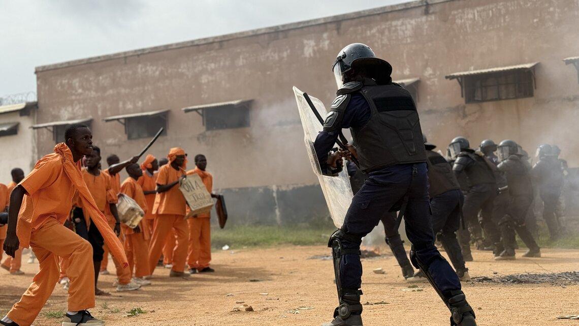 A man in riot police protective equipment standing behind a shield with several prisoners wearing orange uniforms standing front of them.