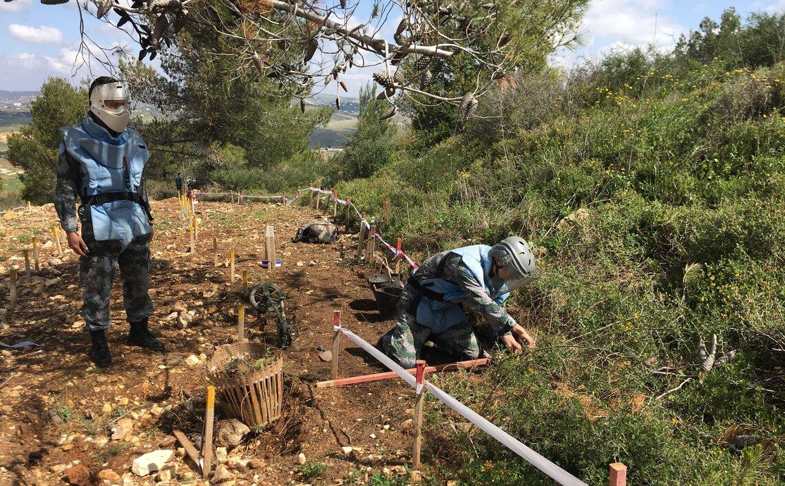 Two peacekeepers in protective gear examine the ground under some trees