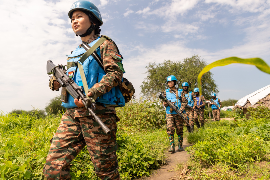 Armed male and female peacekeepers walk in a line between low-lying bushes