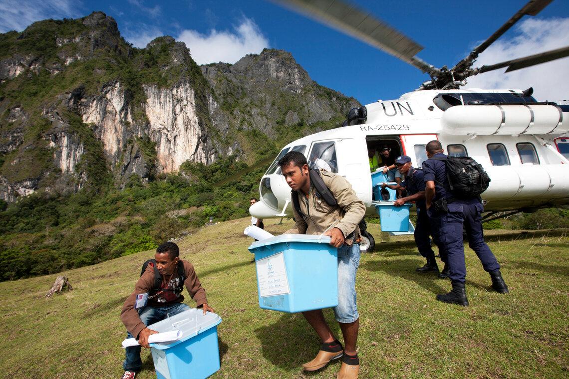 A group of people unload blue plastic containers from a UN-marked helicopter that has landed in a grassy area. Mountains are visible in the background, and the scene appears to depict a logistical operation, possibly involving the delivery of supplies or humanitarian aid.