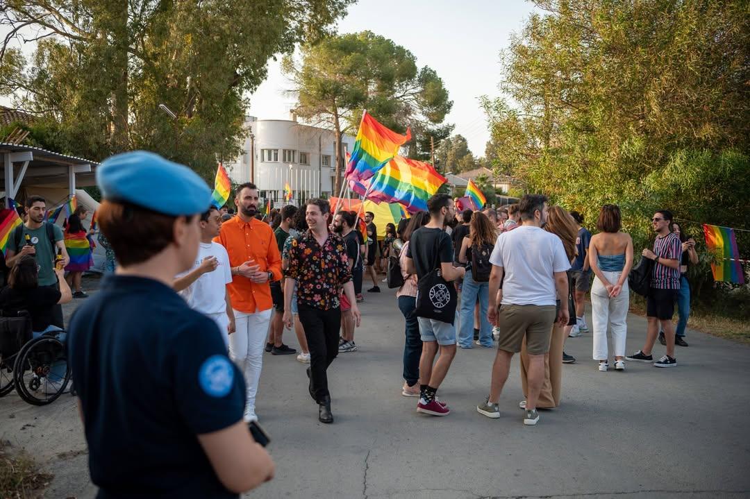 People march in a pride parade waving rainbow flags, while a UN peacekeeper looks on