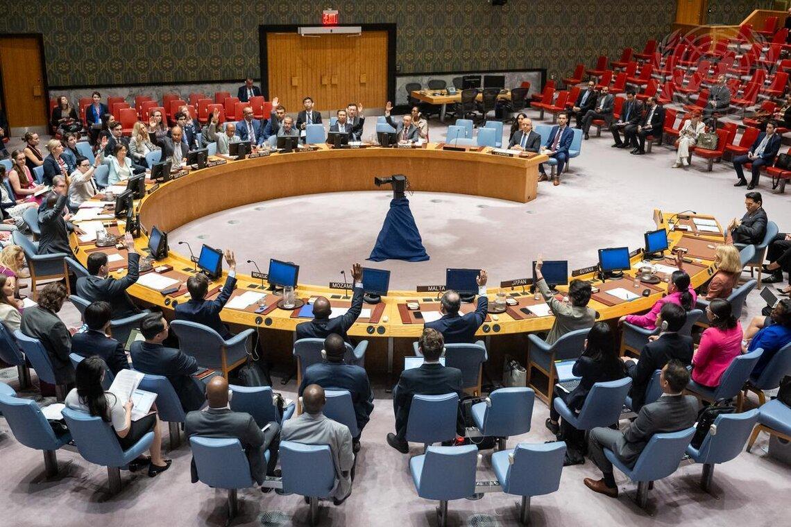 Member State representatives sit in chairs arranged in a circle in a large room, raising their hands