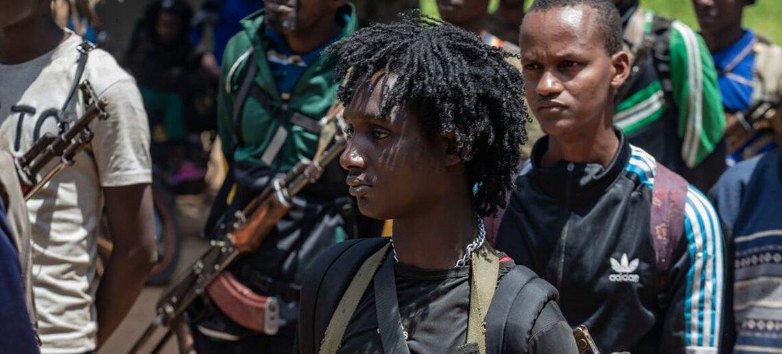 A young man stands in front of a group of men carrying weapons.